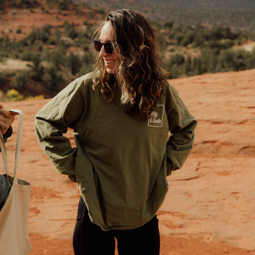 Person wearing an olive green long sleeve with a logo, standing in a desert landscape with mountains in the background.