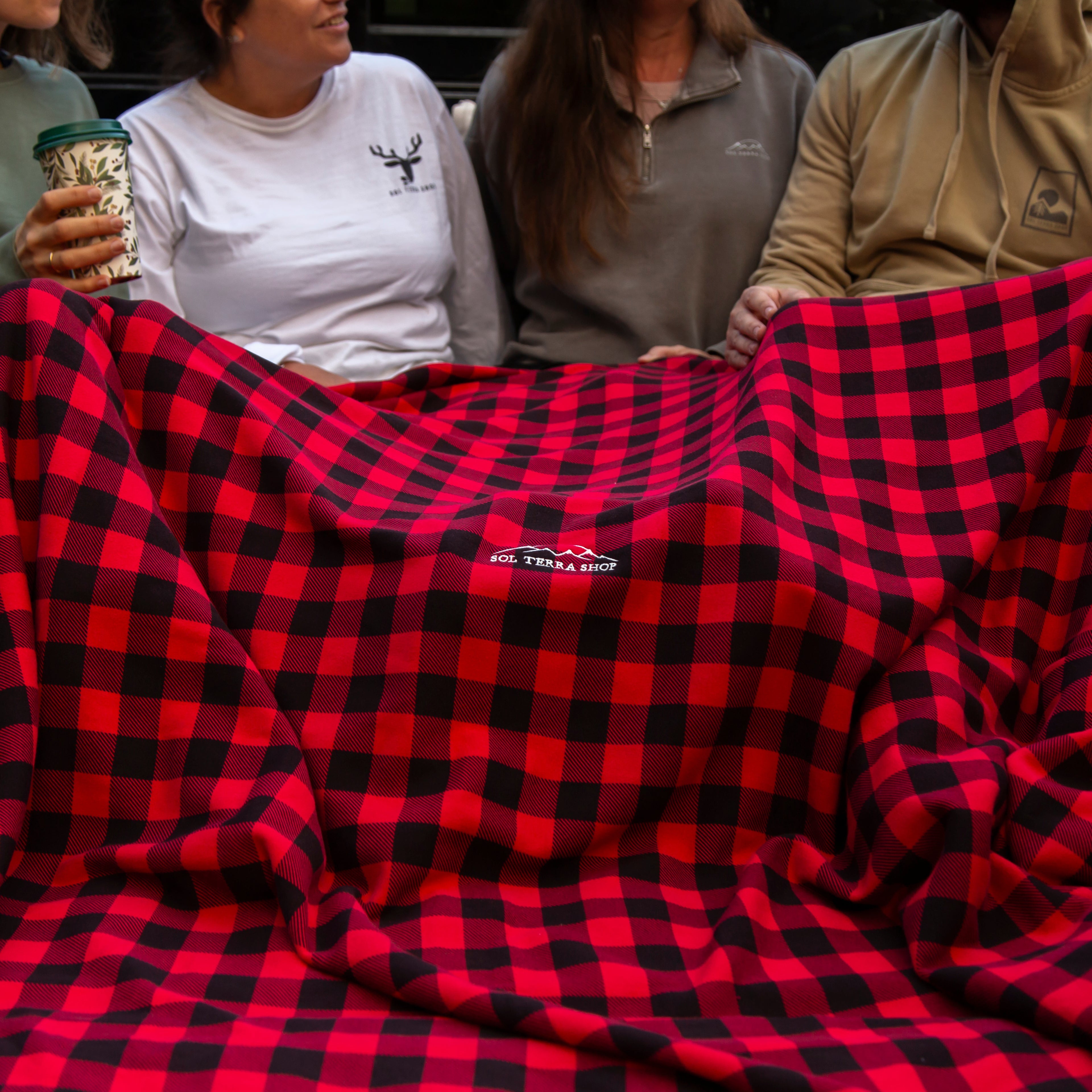 Red and black checkered blanket with Sol Terra Shop logo held by people in a vehicle.