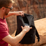 Man in red shirt opening a black recycled backpack outdoors.